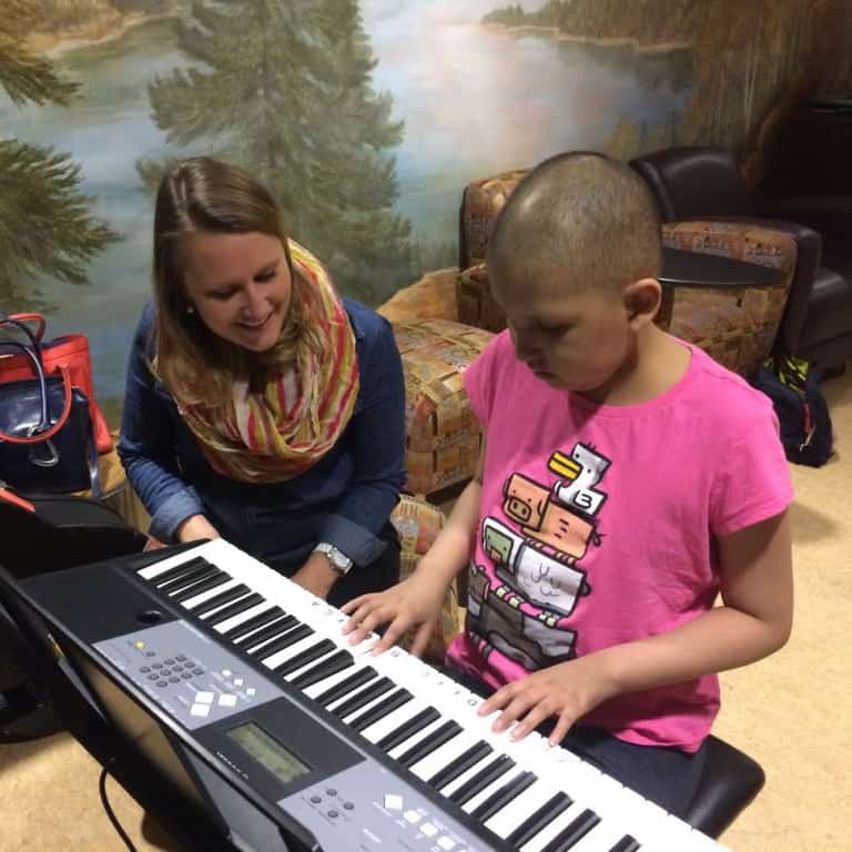 Music therapist Amy Sweetin of Swallow Hill Music teaches an individual music therapy session at Brent's Place in Denver - Amy is a white woman with light brown hair wearing a blue shirt and a yellow scarf, and is smiling as she leans over an electronic keyboard played by a child with buzzed hair and a pink shirt who is looking down at the keyboard.