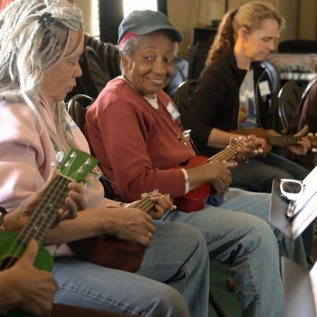 An older black woman playing a ukulele and wearing a hat looks at the camera and smiles during Musical Memories, a group music therapy session at Swallow Hill Music in Denver - two women also playing ukuleles and looking at music stands are to her left and right.