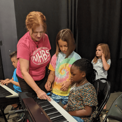 An image of instructor Stef Kull, a woman with red hair, bending over an electric keyboard to teach 3 observing kids how to play the piano during a class at Swallow Hill Music - in the background, another boy concentrates while playing a keyboard.