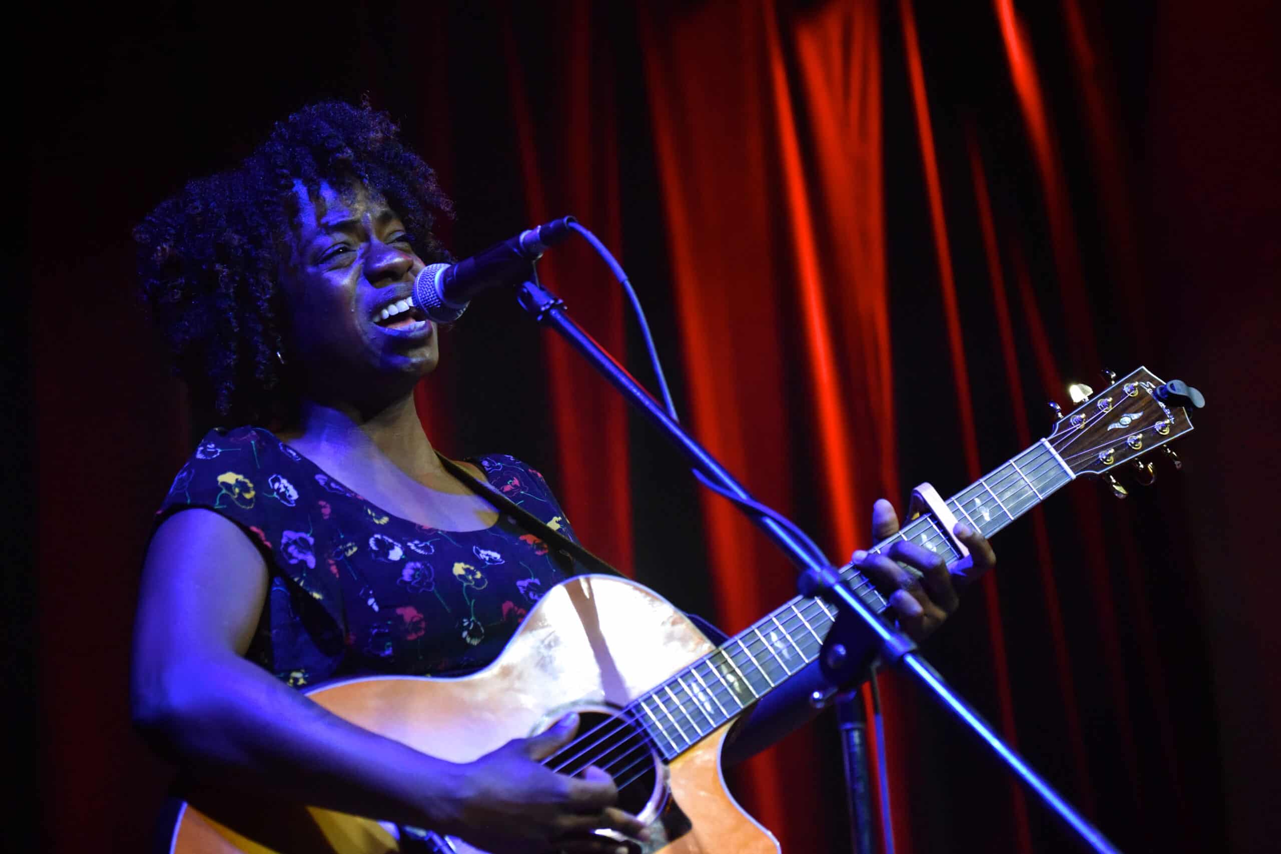 Blues artist Sunny War, a black woman, holds an acoustic guitar and sings into a microphone during a concert at Swallow Hill Music, a nonprofit music venue in Denver.