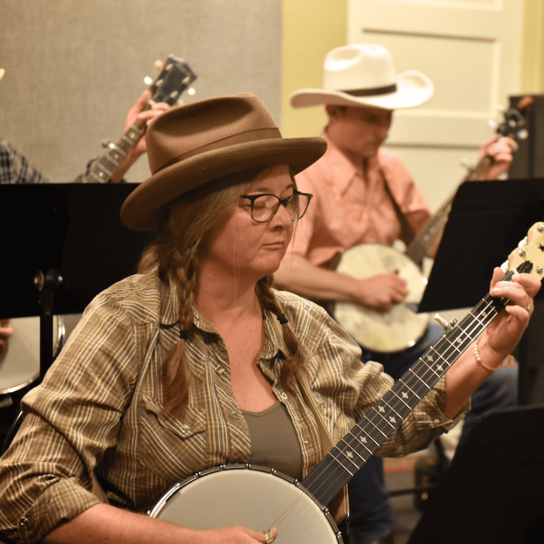 A white woman wearing a cowboy hat and glasses looks down at her music stand while playing the banjo during a music class at Swallow Hill Music.