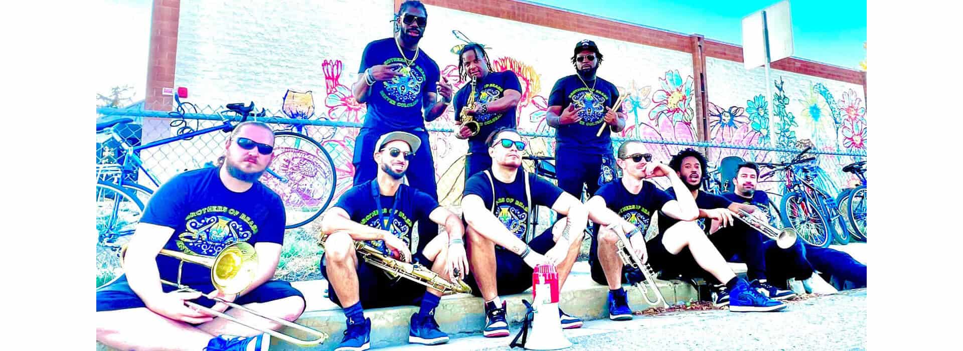 An image of the 9 members of the Brothers of Brass, headlining the 2023 Clyfford Still Lawn Concert produced by Swallow Hill Music, leaning against a wall with various brass instruments