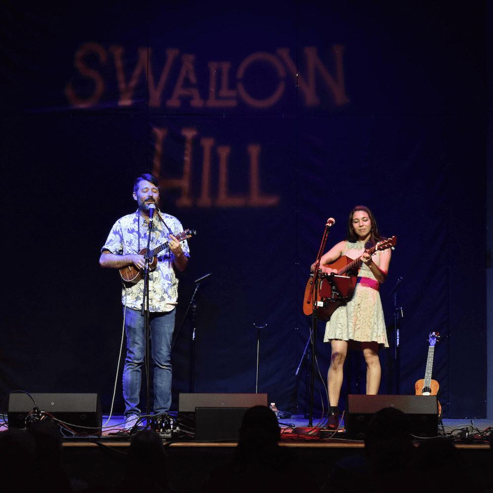 A man playing a ukulele and a woman playing a guitar to his left, both singing, perform onstage in Daniels Hall at Swallow Hill Music with the words "Swallow Hill" projected behind them