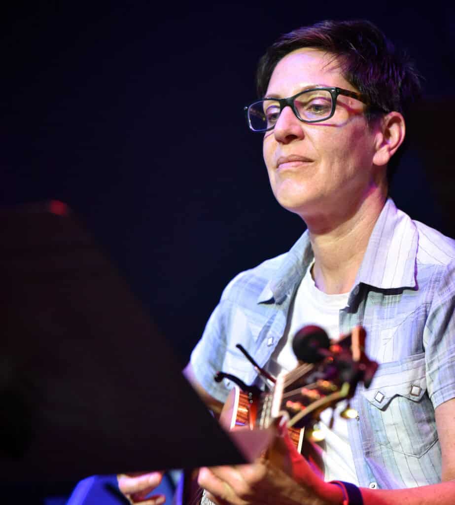 A woman with short dark hair and glasses looks down at a music stand while sitting and playing an acoustic guitar, slightly smiling