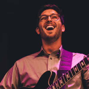 Thomas Jennings, the instructor for Swallow Hill's Progressions: Electric Guitar online course, is a man with black glasses and short black hair. He is playing a large hollowbody electric guitar while looking away from the camera and smiling.