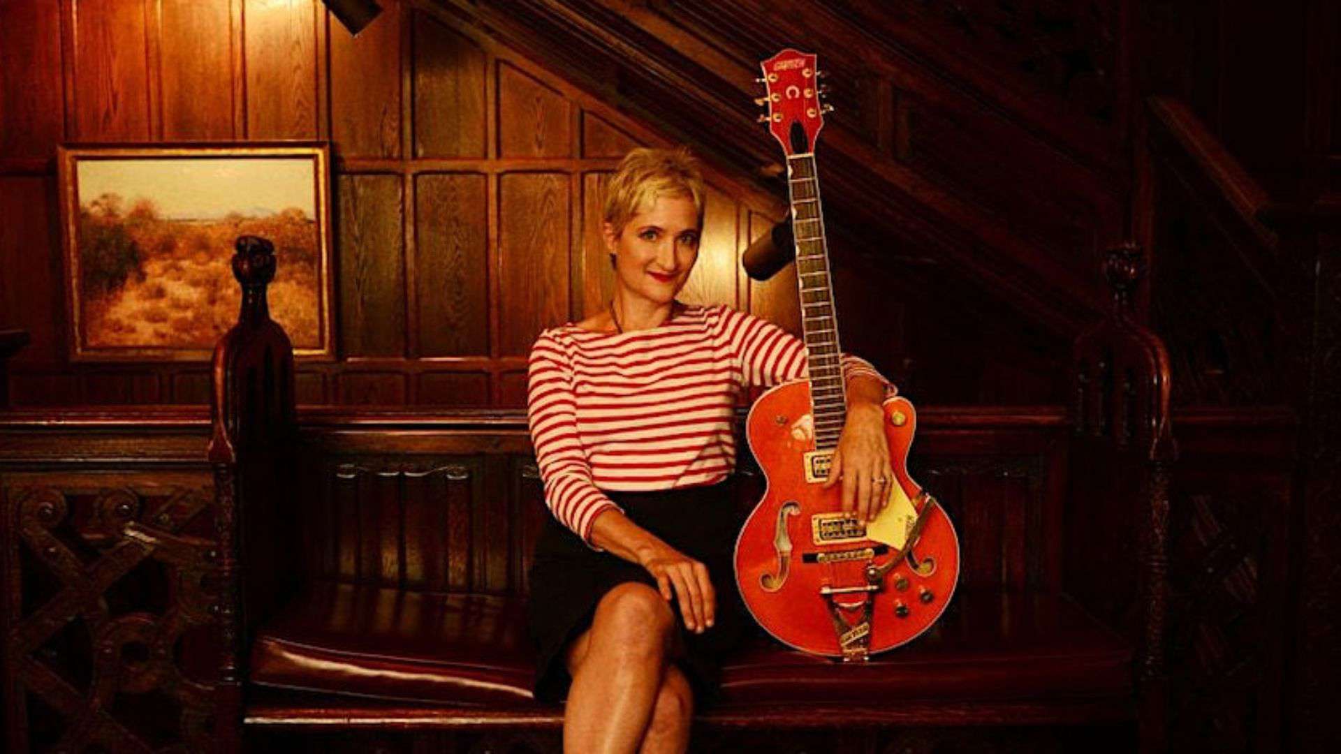 A headshot of artist Jill Sobule performing at Swallow Hill Music, a white woman with short blonde hair sitting on a couch, holding a large hollowbody guitar in her right hand and wearing a striped shirt.