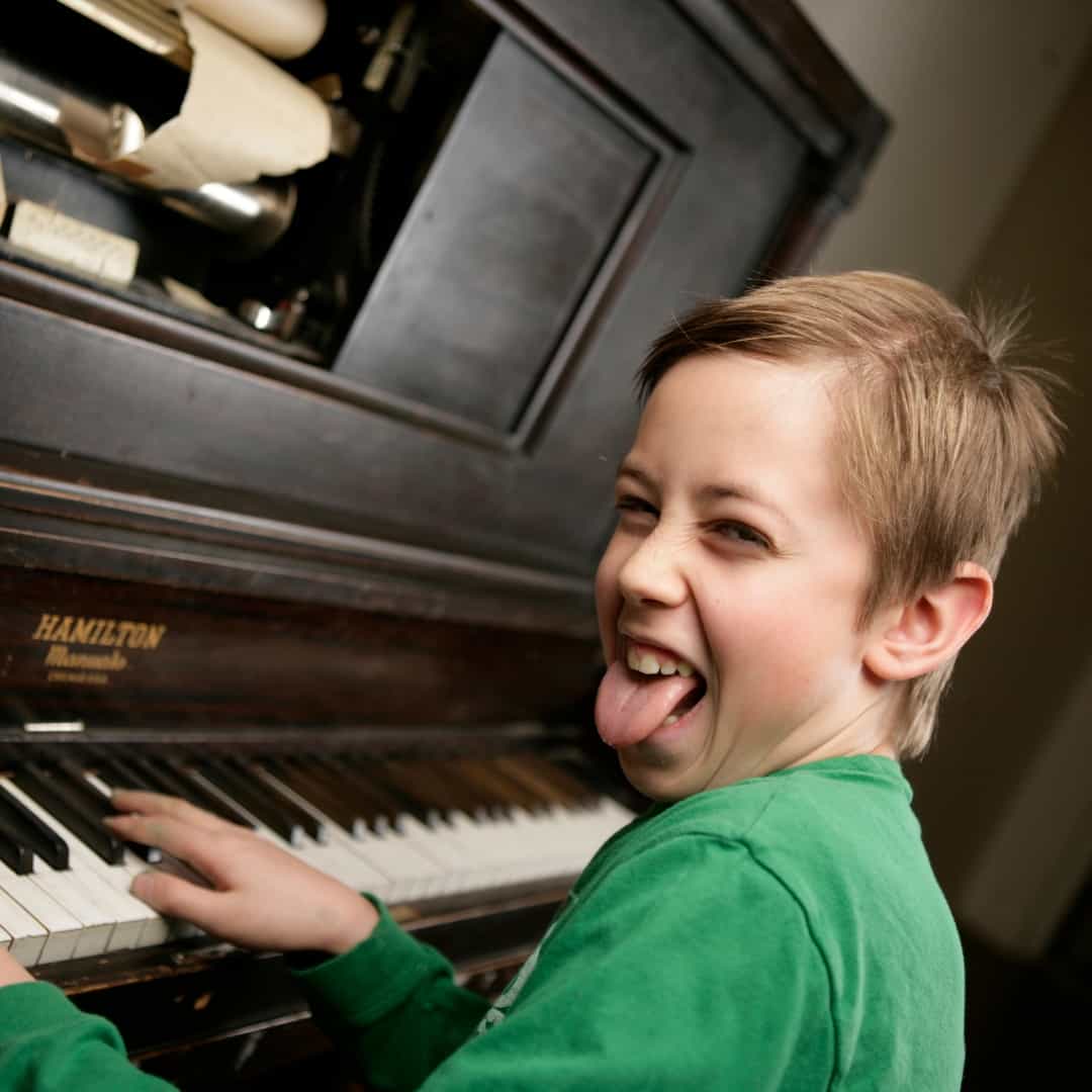A young boy sticking out his tongue looks at the camera while playing the piano