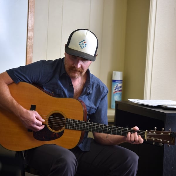A photo of Jarett Mason, a music instructor at Swallow Hill Music, sitting and bent over an acoustic guitar he is playing and looking down at.