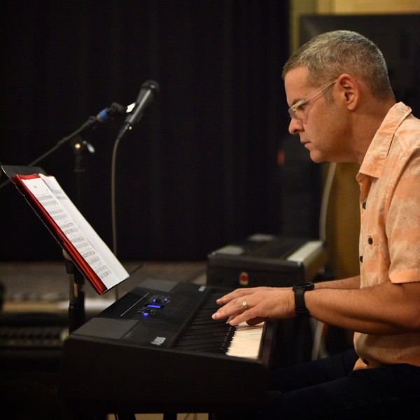 A photograph of a man with glasses seated and looking at sheet music on a music stand while playing an electric keyboard.