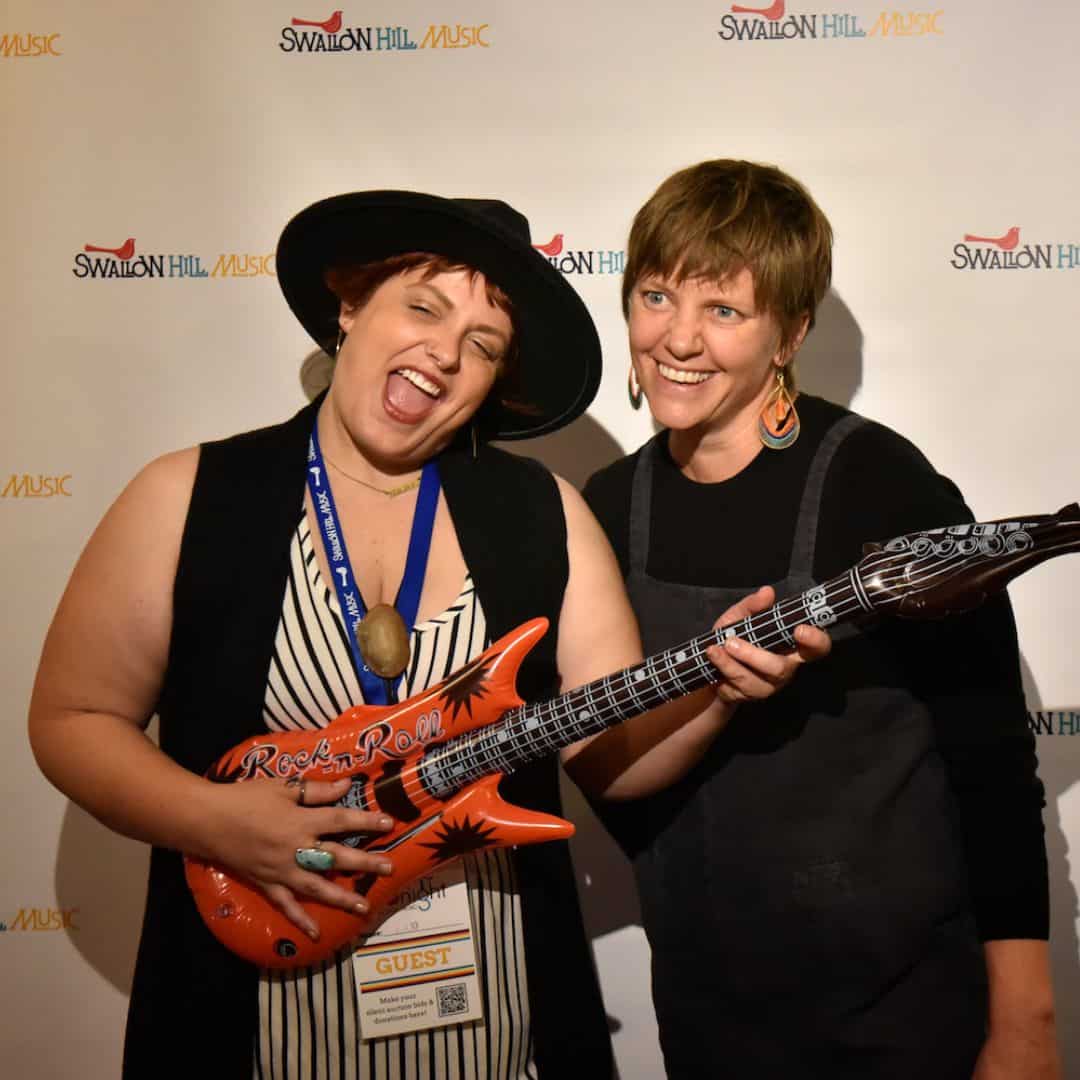 Two people stand in front of a photo backdrop with the Swallow Hill Music logo on it and pose for a picture - the person to the left is making an excited face and playing a blow-up toy electric guitar
