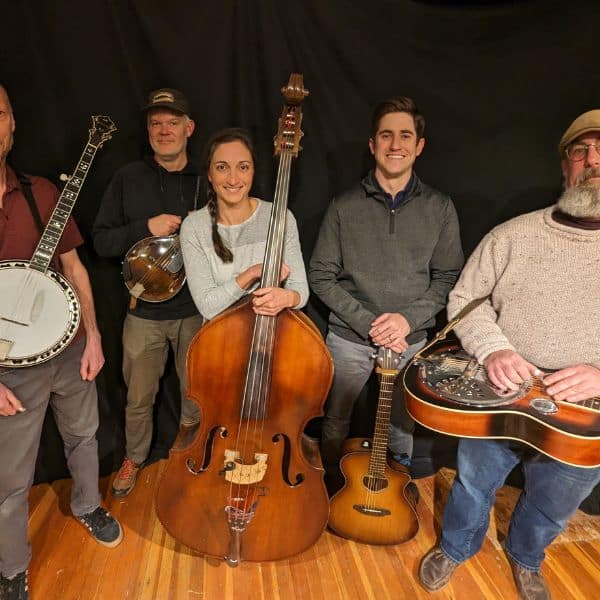 Four men and a woman in the center all face the camera and smile holding instruments; from left: banjo, mandolin, upright bass, guitar, and dobro.