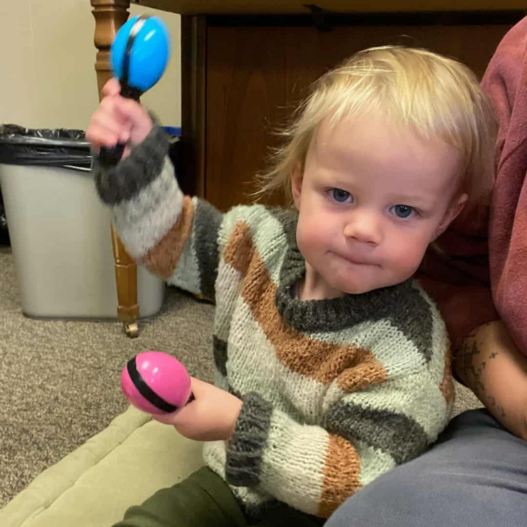 A blonde toddler shakes maracas while looking into the camera and sitting during a music class at Swallow Hill Music in Denver.