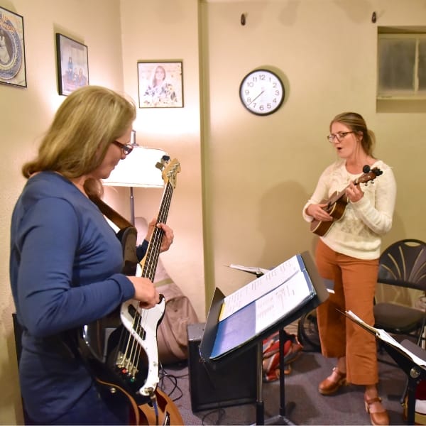 A standing woman plays bass guitar while looking down at a music stand, and to the right of her another woman stands and plays the ukulele and sings in a classroom with a clock and concert posters on the wall during a music class at Swallow Hill Music in Denver.