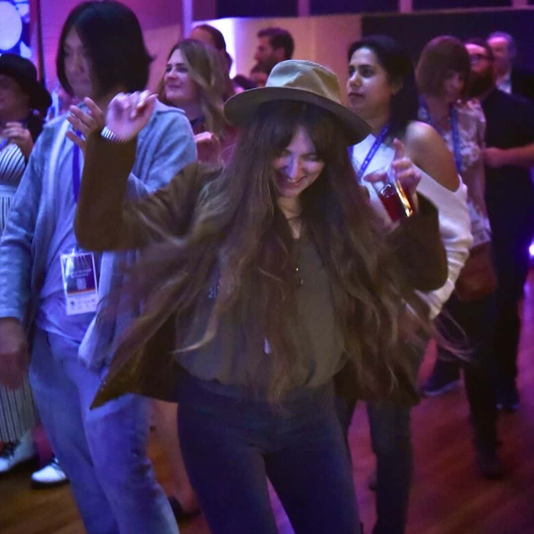A young woman in Daniels Hall at Swallow Hill Music with long hair wearing a hat dances and smiles with her arms raised.