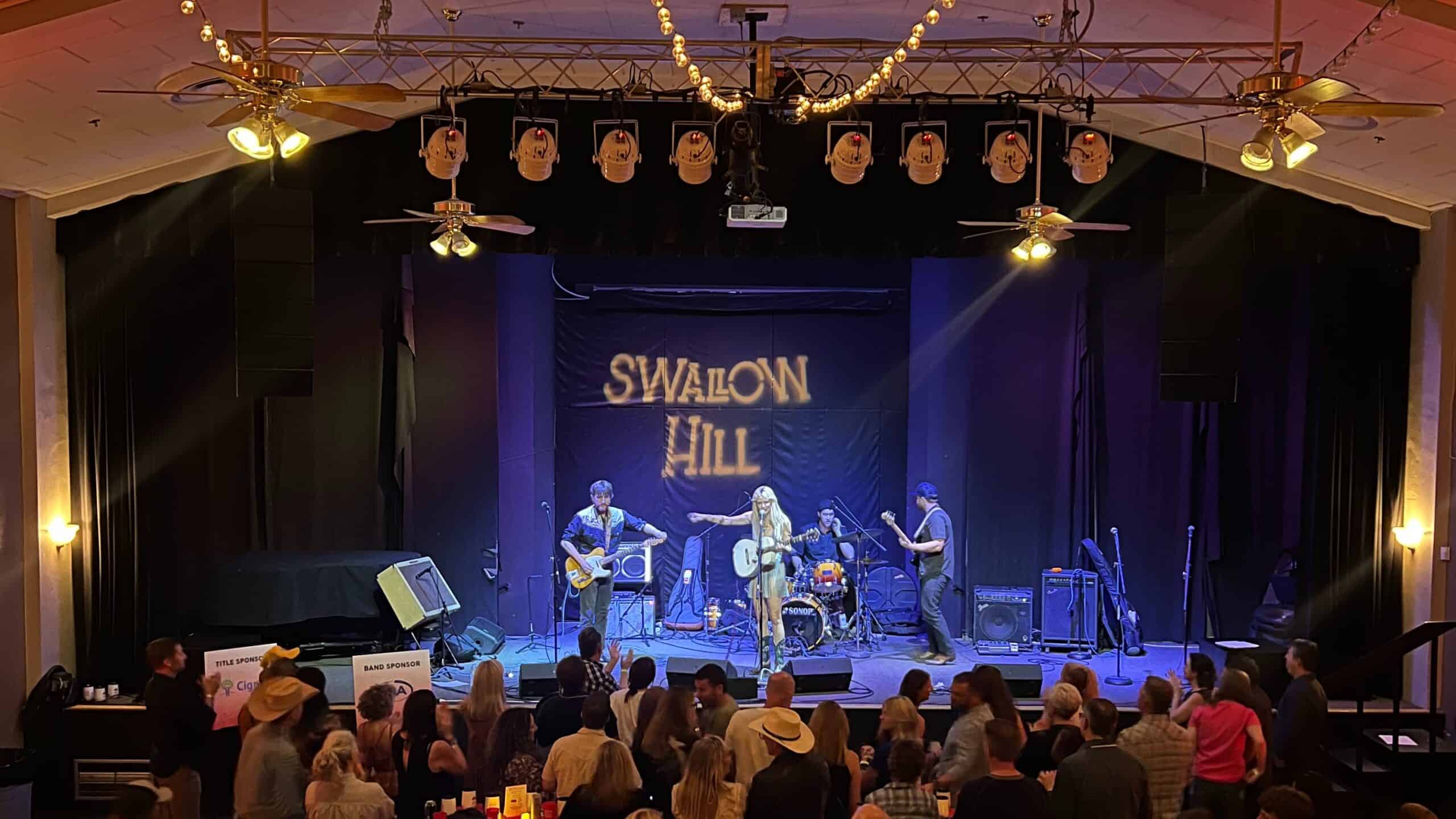 A photo of a crowd in the foreground watching a concert on the stage at Daniels Hall at Swallow Hill Music, with a black curtain behind a band and the words "Swallow Hill" projected upon the curtain.