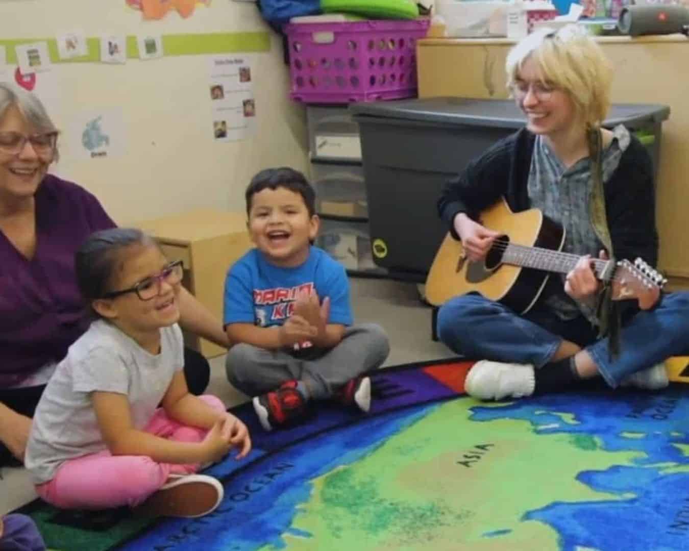 A woman with short blonde hair and glasses smiles while playing the guitar and singing, seated on a floor with two preschoolers and their teacher at a Little Swallows music class presented by Swallow Hill Music in Denver.