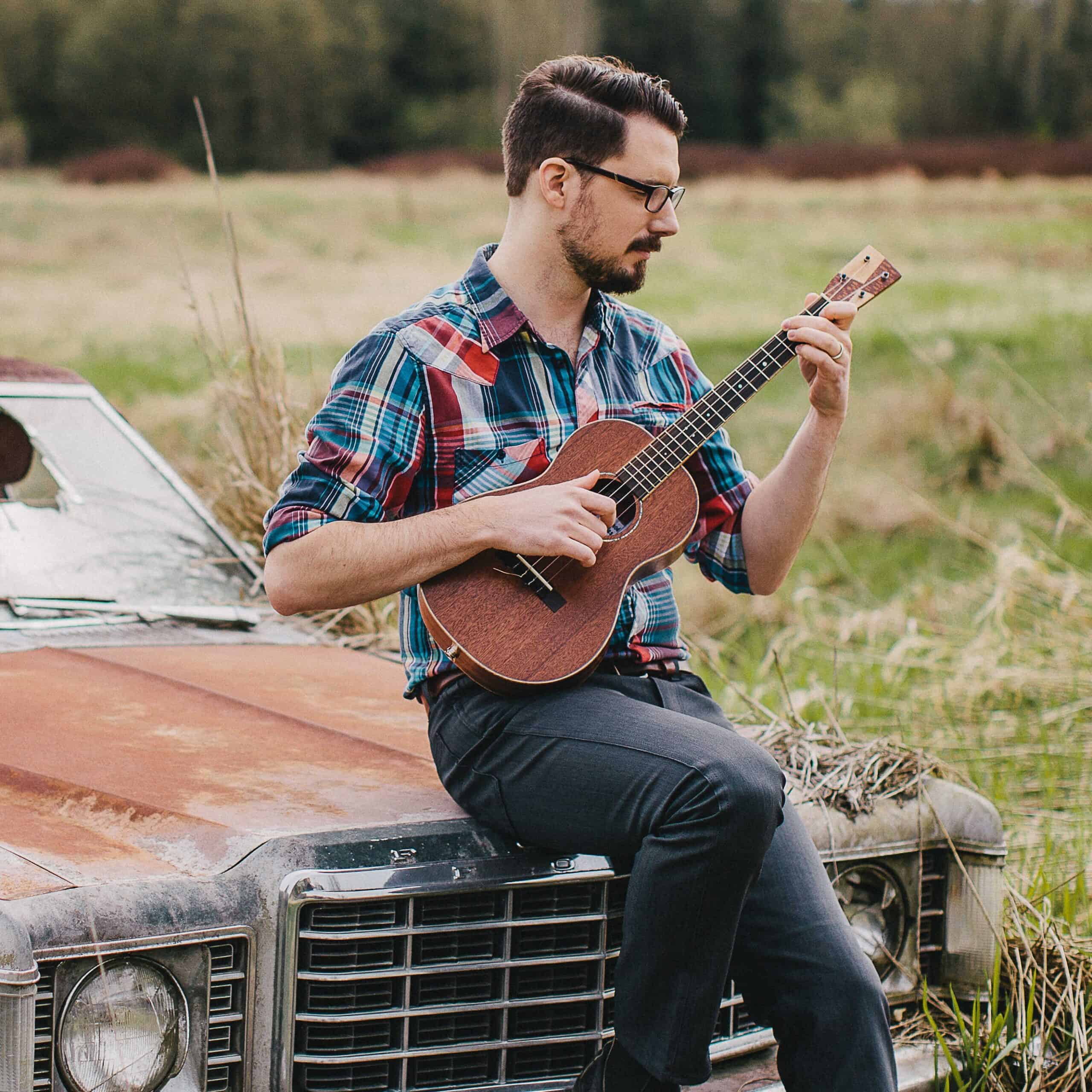 Denver Ukefest 2024 Friday Headliner James Hill, seated on the hood of an old car playing his uke