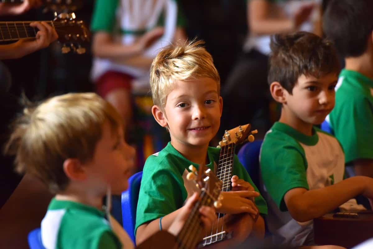 A young blonde boy looks at the camera and smiles while holding a ukulele, with two boys looking elsewhere also holding ukuleles on his left and right, during an Explore! Music Summer Day Camp at Swallow Hill Music.