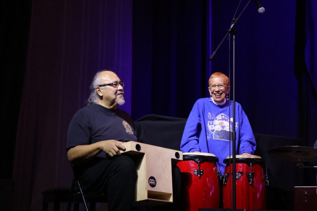 An older female student and instructor Ed Contreras play hand percussion drums while smiling onstage during a class at Swallow Hill Music in Denver.