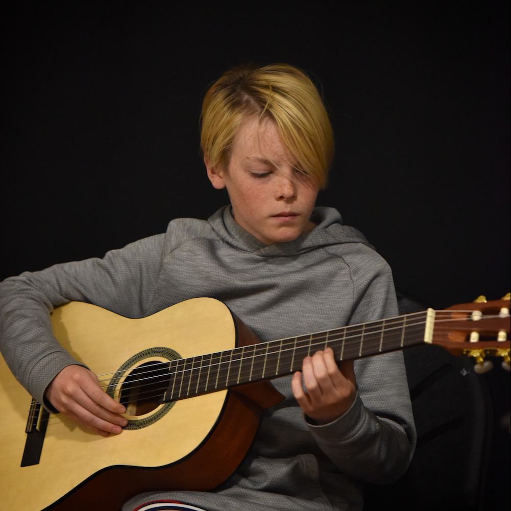 A young boy looks down at an acoustic guitar while playing it
