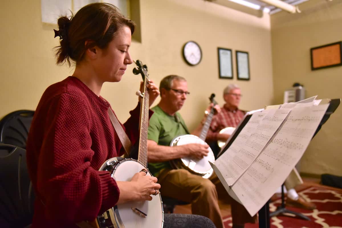 A young woman looks down at a music stand while playing a banjo in a group class at Swallow Hill Music in Denver - several other banjo students behind her do the same.