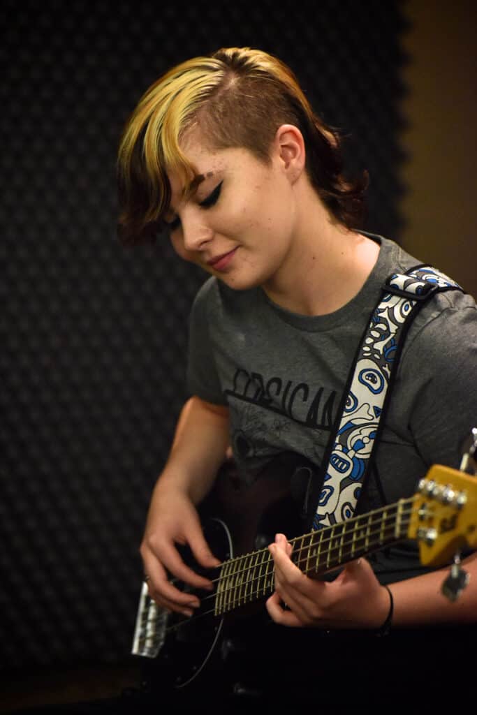 A teen student smiling while they look down at a bass guitar they're playing during a class at Swallow Hill Music in Denver
