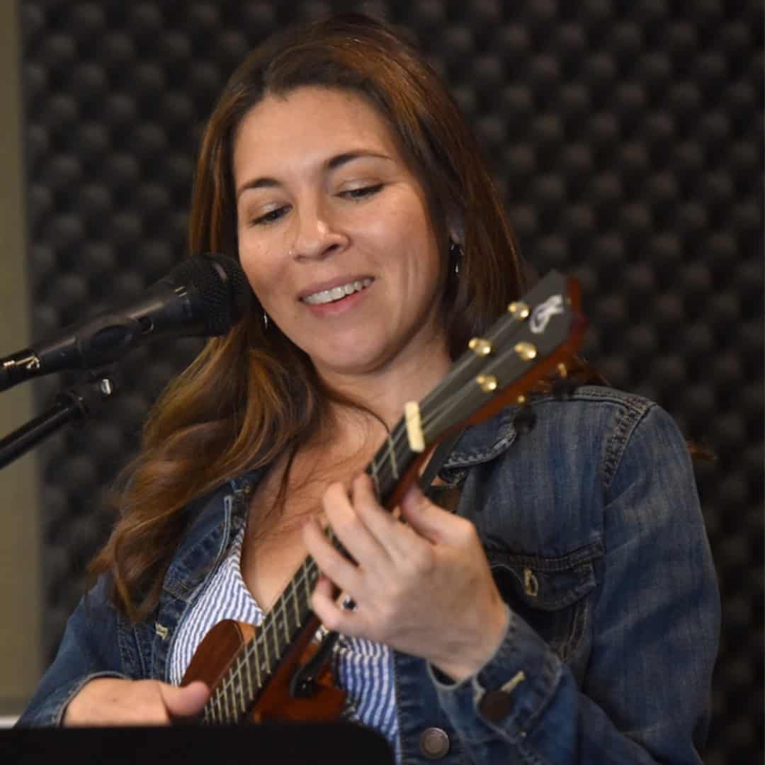 A woman looks down at a ukulele while playing it in front of a microphone while teaching a Ukulele class