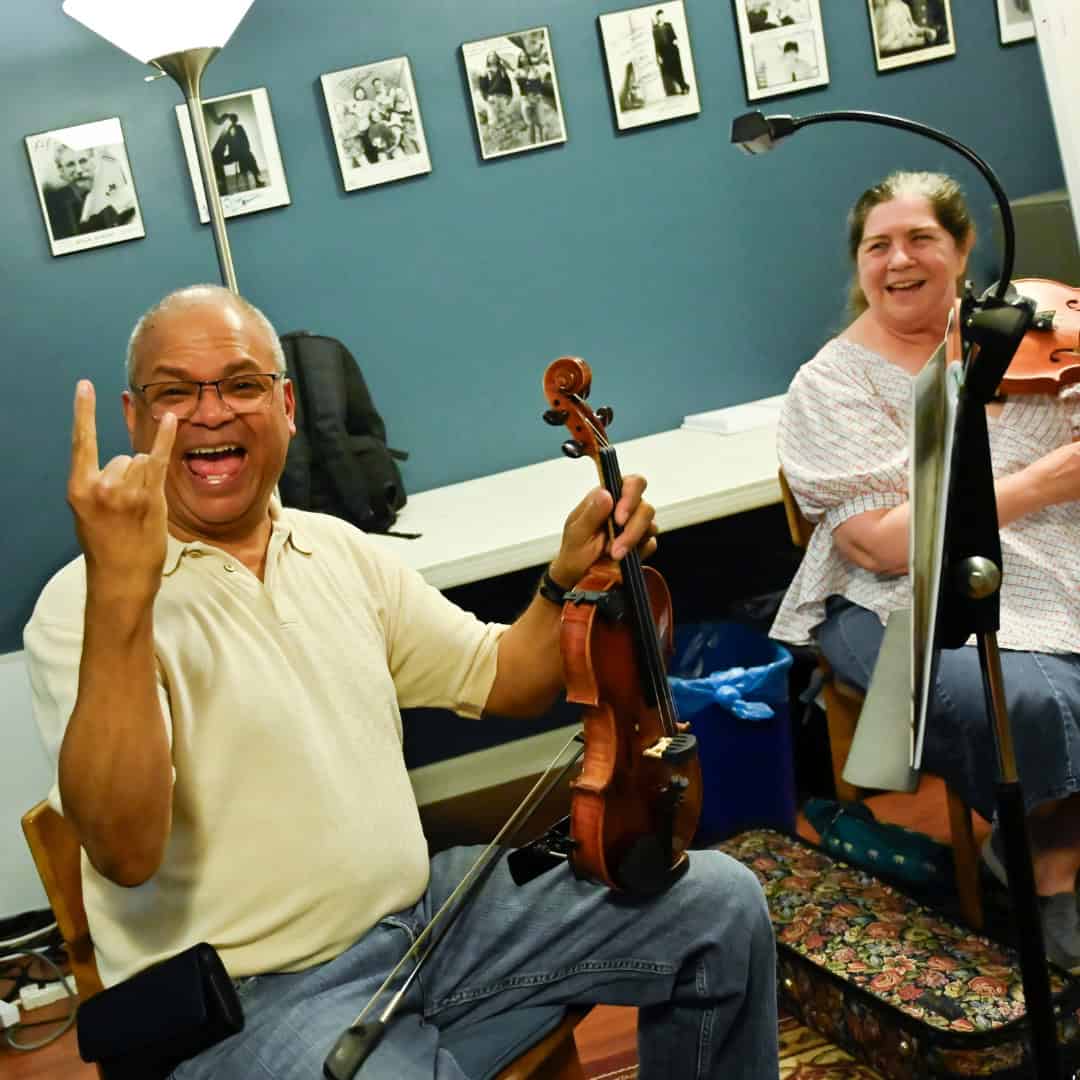 A smiling man with glasses holding a violin looks into the camera and smiles and throws the horns while holding a violin on his knee during a music class