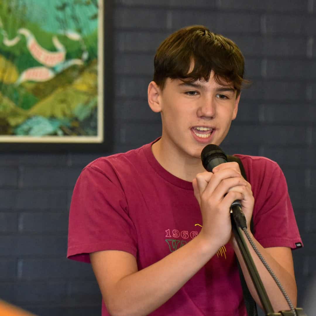 A teen boy looks down at an electric guitar he is playing in front of a microphone stand and a music stand