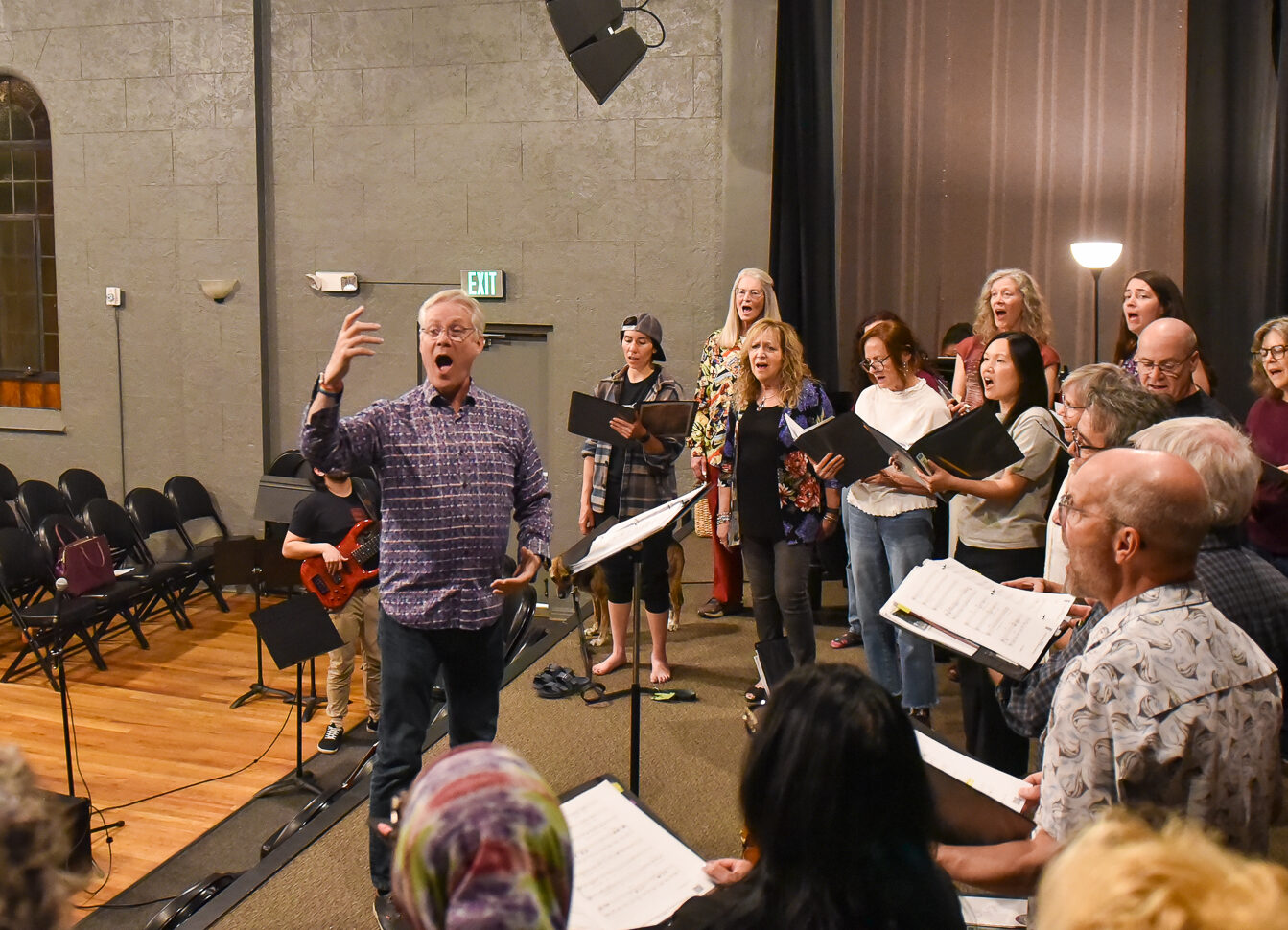 A man directs a choir of around 30 people of various genders and ages with his hands on stage in a concert hall while singing