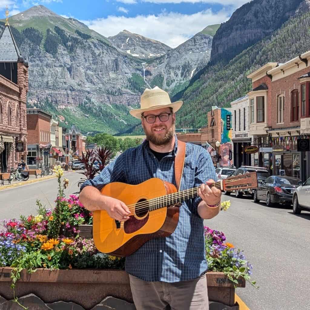 An image of Martin Gilmore, instructor of the Progressions: Songwriting course, standing holding a guitar by a flower bed on a street with mountains in the background