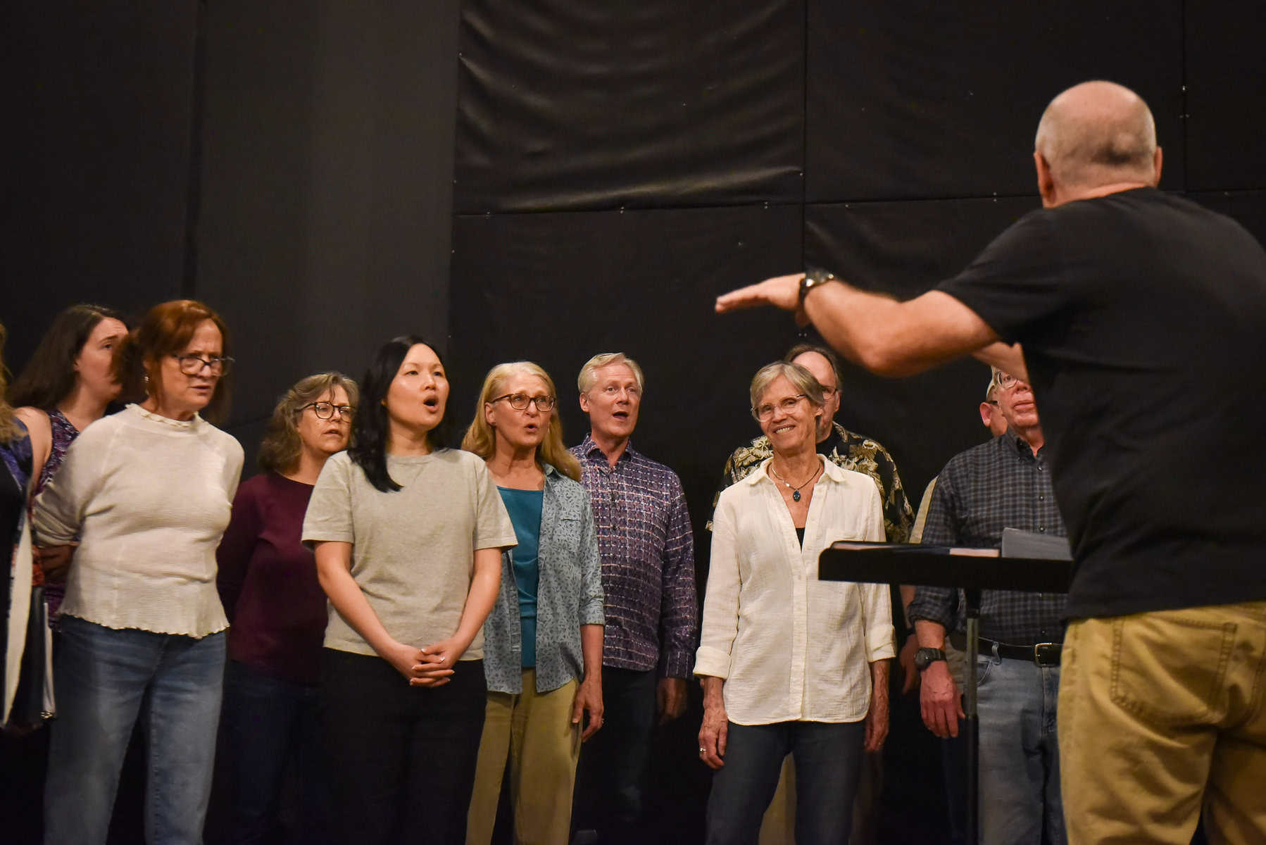 Kelley Contreras Memorial Choir mixed gender group Roy conducting A photo of a man in the foreground from behind raising his arms while conducting a portion of a choir of various ages and genders, who are facing the camera singing