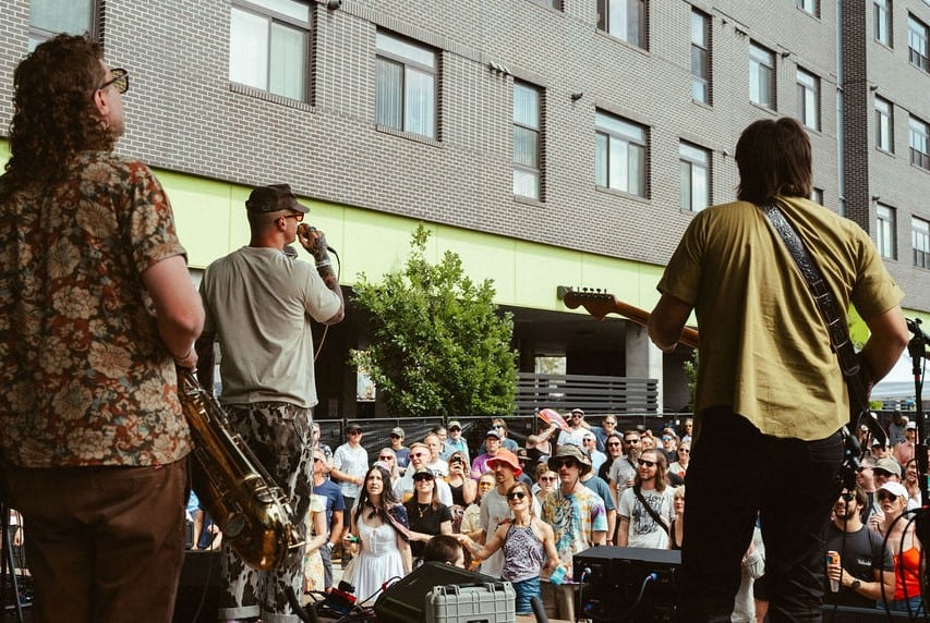 Indiewood Rootbeer Richie from behind with full crowd additional shot A photo of a band playing on a stage at an outdoor music festival, taken from behind facing towards a large crowd in front of the stage