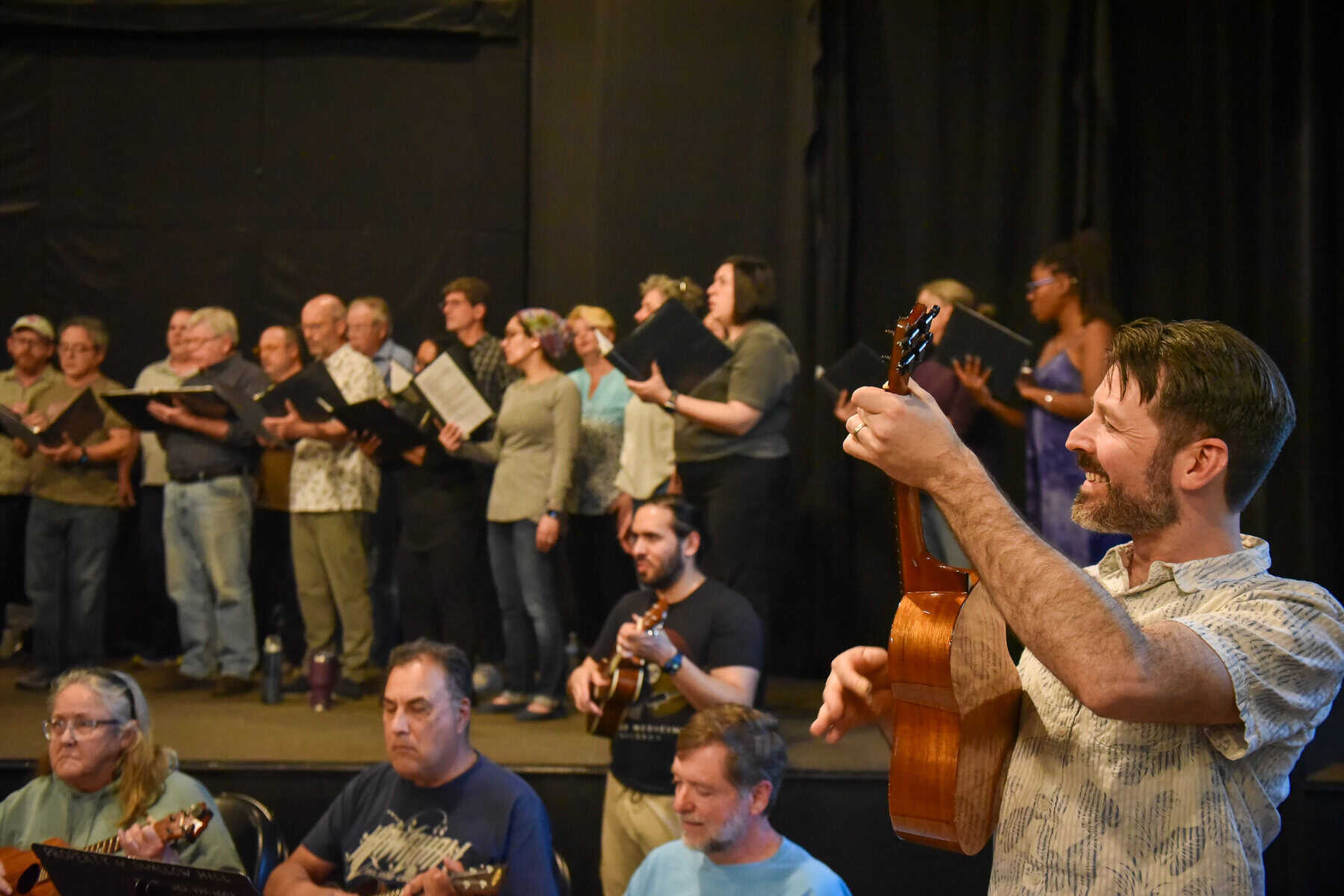 A music teacher with a ukulele in the foreground leads a class full of ukulele players and singers in the background on a stage