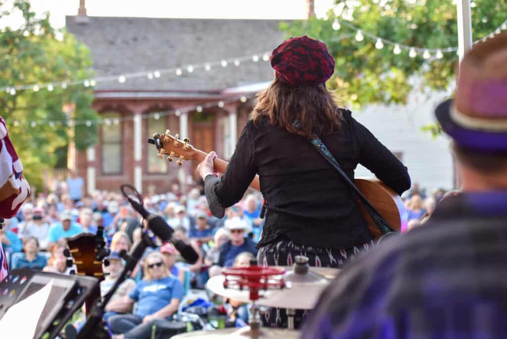 A shot of a woman singing and playing guitar on a stage from behind with a seated audience under string lights visible in the background