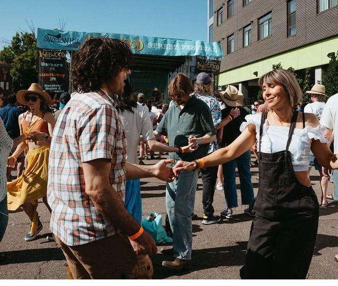 Two adults doing a partner dance during an outdoor music festival with a stage visible in the background