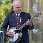 A photo of Pete Wernick standing and playing the banjo wearing a suit while performing at a concert.