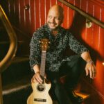 Denver UkeFest performer Steven Espaniola sits on a staircase smiling and holding his ukulele
