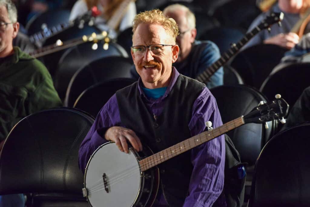 A woman looks at a banjo she is playing while seated with several other banjo players behind her during a workshop at Swallow Hill BanjoFest