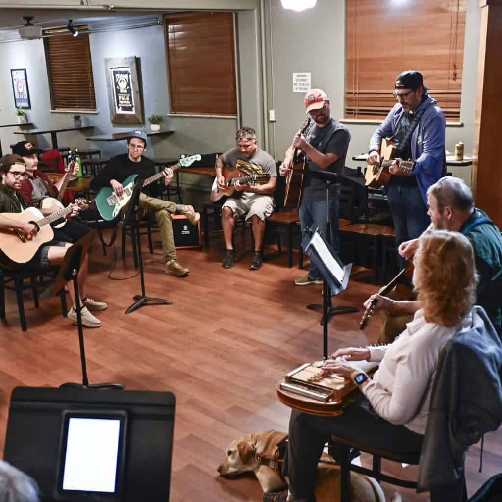 Four men and a woman in the center all face the camera and smile holding instruments; from left: banjo, mandolin, upright bass, guitar, and dobro.